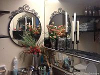 Photo showing two black metal framed circular mirrors with scrollwork, artificial florals, various bottles, candles, and black metal twig candle holder with three candle cups on bathroom counter.