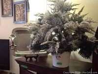 The faux snow-flocked pine plant with large artificial pine cones in a white ceramic vase, on display with the tray visible behind the plant.