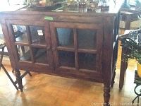Front view of dark brown wood cabinet with two glass-paneled doors and turned legs on parquet floor.