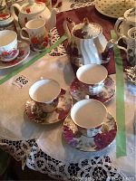 Three teacups with saucers and a teapot arranged on a table with lace tablecloth and placemats.