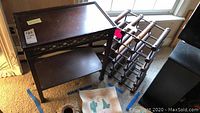 Photo showing wood side table with edged top, a lower shelf and decorative cut-out detail on the top apron, dark wood finish. Adjacent wood wine rack with multiple horizontal slots for bottles, dark wood finish.
