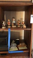 Wooden shelf displaying four porcelain Goebel Hummel figures and additional collectibles below including lions and kaleidoscope.