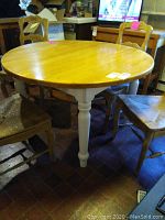 Photo of round wooden kitchen table with light wood top and white painted turned legs, showing the table with no leaf extended and 3 chairs partially visible.