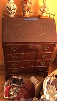 Front view of vintage wooden secretary desk showing carved leaf scroll designs on the drop-front panel and four drawers with ornate brass handles and locks.