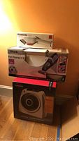 Stacked boxed appliances: Rowenta dress fit steam brush on top, Shark Pet Perfect II vacuum in the middle, Vornado whole room heater on bottom against wall.