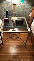 Photo of one glass and metal side table with digital clocks and lamp on top.