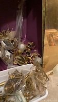 Assortment of brass door handles, knobs, curtain holders, and a brass light fixture with frosted glass shades piled together against a purple backdrop and a mirror.