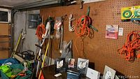 Wide view of garage corner with various tools and extension cords hanging on pegboard and standing against wall; some framed photos not included.