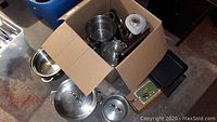 Photo showing a cardboard box containing various stainless steel cookware items, including pots, a steamer insert, and lids, and a Cuisinart blender base with pitcher.