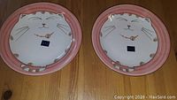 Two round ceramic plates with pink edges and a white kitten face design on top.