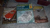 Photo displaying three older automotive books placed on a carpeted floor: Science and Mechanics guide for electronic garage door openers, Oldsmobile Cutlass automotive repair manual (1974-1988), and a Mechanix Illustrated magazine dated 1955.