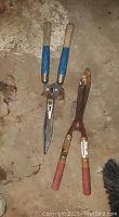 Photo shows two manual hedge trimmers with visible rust and wear, one with blue handles and the other with red handles.