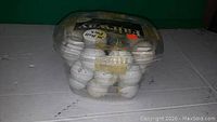 Clear plastic container holding about 25 used white golf balls with various markings, some scuffed or dirty.