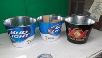 Photo showing three metal beer cooler buckets, two blue with Bud Light logo and one black with Dos Equis logo on a white surface.