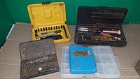 Photo showing six tool kits and storage cases including screwdriver bit set, hand tools in metal boxes, and plastic organizers.