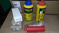 Photo showing two yellow bottles of blue and red chalk fluid, a red chalk line reel, and several boxes of fasteners.