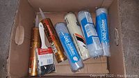 Cardboard box containing multiple paint rollers, liquid nail tubes, and paint brushes.