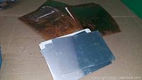 Six metal sheets of 8 inch size, two copper sheets with patina and four aluminum sheets with smooth finish, placed on cardboard on floor.