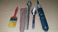 Photo showing a yellow bristled paint brush, metal ratchet wrench with fabric pouch, metal spoon with black handle, and a blue electric tool with textured grip placed on cardboard surface.