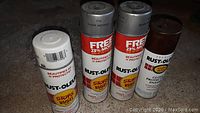 Four Rust-Oleum spray paint cans in different colors: white, two silver metallic, and brown, placed on the floor.