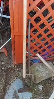 Photo of a pitch fork and garden hoe leaning against an orange lattice panel and other miscellaneous outdoor items on the ground.