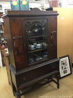 Full view of antique wooden china cabinet showing glass door with wooden scroll design and single drawer below.