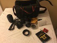 Image showing Minolta X-370 camera, additional lens, flash, lens hood, camera bag, manuals, and other accessories on a white cloth.