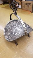 Metal wood caddy angled to show embossed floral and geometric pattern on tray, arched handle with decorative knob, and legs