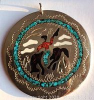 Front view of round silver-toned pendant with turquoise and coral inlays showing cowboy riding bucking bronco motif and decorative scalloped edge