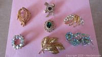 Top view of 7 vintage gold-tone Coro brooches on pink background showing different designs with rhinestones and textured metalwork.