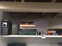 Shelf displaying metal ammo box, stack of military books including Camp X and A Man Called Intrepid, and Boutell's Heraldry book.
