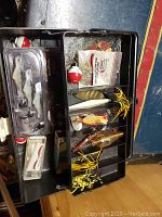 Open metal fishing tackle box showing compartments with fishing flies, bobbers, and fishing accessories.