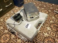 Photo showing two gray plastic cases stacked with an Otari power supply and a circular electrical component on top, placed on a patterned carpet.