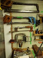 Various hack saws and hand saws hung on pegboard with several tools including wrenches and hammers on shelf below.