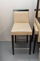Front-facing view of beige upholstered counter stool with dark wood legs and metal footrest on bottom.