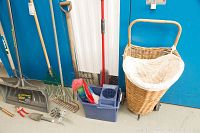 Photo showing antique wicker laundry basket on wheels, multiple garden rakes, pitchfork, snow shovels, and cleaning tools composed along blue wall.