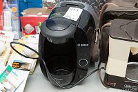 Photo shows a black Bosch Tassimo coffee maker with front pod compartment and cord extended, next to a Melitta drip coffee maker and assorted light bulbs.