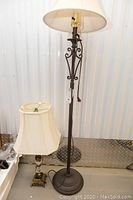 Pair of lamps including one floor lamp and one table lamp shown together. Tall lamp measures 57 inches and has a beige fabric shade, decorative metal base with scroll details. Smaller lamp has a cream narrow fabric shade and square metal base.