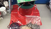 Red Christmas tree stand with green top and metal clamps, bag with black cable wire and other small items, roll of wrapping paper, and packaged Christmas ornament on blue background.