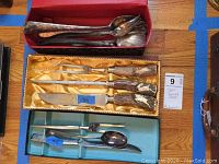 Full view showing the Klaus Tragbar carving set in box with antler handles and large silver serving spoons in red box above and other serving utensils below.