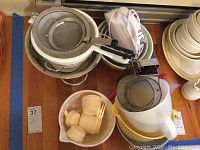 Kitchen lot showing stacked metal colanders, mixing bowls, plastic measuring cups in a cream yellow, and a white electric hand mixer on top.