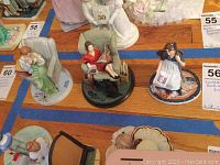 Three Norman Rockwell figurines including woman reading, girl with dog holding magazine, and woman with baby in rocking chair.