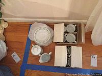 Photo showing boxed tea cups and saucers along with creamer and sugar bowl grouped on wooden floor