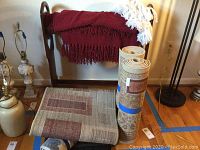 Photo showing wooden blanket rack with dark red and white fringed blankets draped over it, two rolled area rugs standing upright, and two area runners on the floor.