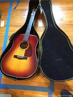 Full view of the Fender sunburst acoustic guitar resting inside an open black hard case with yellow trim, showing wood grain floor underneath.