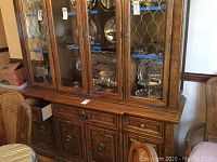 Full frontal view of the china cabinet showing glass display upper section with silver plates and other items inside, wood lower section with drawers and cabinets.