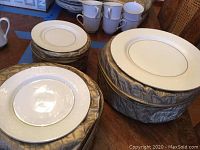 Photo showing stacks of white Mikasa Bridal Veil pattern plates with gold rim in zippered quilted storage cases along with some cups in background