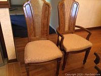 Two high back wooden dining chairs side by side. One chair has armrests, both have cane inset backs and beige cushioned seats.