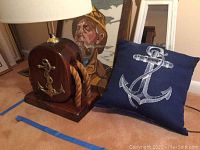 Photograph shows dark wood nautical lamp with rope and brass anchor, blue pillow with white anchor print, and framed fisherman artwork partially visible behind lamp.