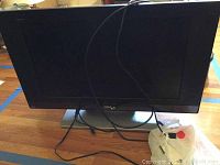 Photo showing the front of the black RCA flat screen TV from a slight angle, with visible cables and part of the base stand on a wooden floor.